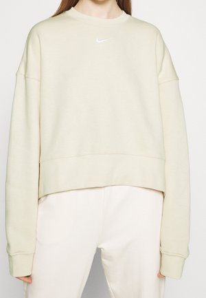 Femme portant un sweat-shirt Nike beige clair surdimensionné et un pantalon assorti beige clair, sur fond uni.