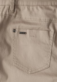 Beżowa tkanina o gładkiej fakturze ma tylną kieszeń z metalicznym akcentem oraz małą metkę z logo. Sutki są widoczne wzdłuż krawędzi.