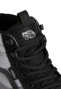 Primer plano de unas zapatillas Vans negras y grises con cordones trenzados, ojales de metal y el logo "Vans Off The Wall" en la lengüeta y en la etiqueta lateral.
