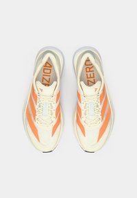 Helle cremefarbene Sportschuhe mit orangefarbenen Akzenten, Obermaterial aus strukturiertem Mesh, Design mit drei Streifen und gedämpfter Sohle. Markenlogo auf der Zunge sichtbar.