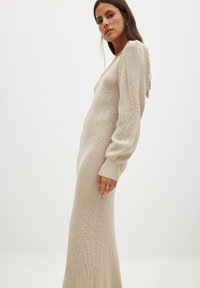 Beige stickad klänning med långa, åtsittande ärmar, en v-ringning och en ribbad textur. Kjolen sträcker sig ner till anklarna, vilket skapar en strömlinjeformad siluett.