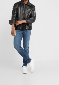 Chaqueta de cuero negra con cierre de cremallera y hombreras, combinada con jeans de mezclilla azules y zapatillas blancas.