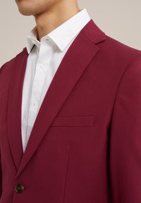 Burgundi spalvos švarkas su lygiu paviršiumi, užsegamas vienu mygtuku, su įpjovomis ant apykaklės ir krūtinės kišeniu, dėvimas ant balto marškinėlių.