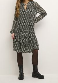 Robe vert foncé avec un motif ondulé blanc, manches longues, décolleté en trou de serrure et ourlet à volants. Associée à des bottes noires à semelles épaisses.