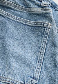 Denim bikses ar gaiši zilu tonis, ar kontrastējošām šuvēm un kabatas detaļu. Audumam ir gluda tekstūra ar smalku rakstu.