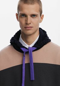 Sudadera con capucha que presenta un diseño a bloques de color en marrón claro, negro y con un acento de lazo púrpura; textura suave y tela blanda.