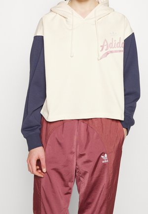 Persoon draagt een crème-witte en marineblauwe hoodie met de tekst "Adidas" en bordeauxrode Adidas trainingsbroek met logo op de dij, staand tegen een witte achtergrond.