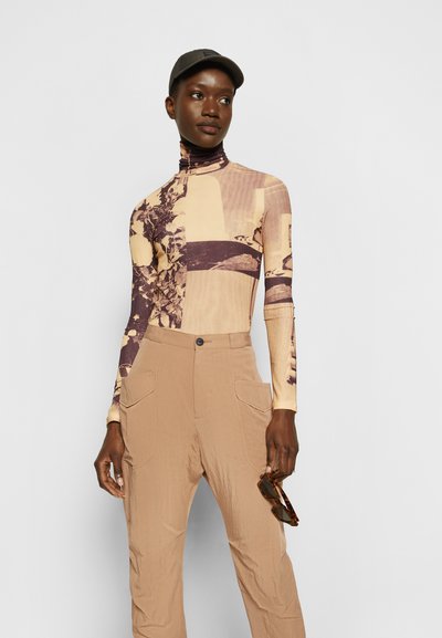 Camisola de manga longa com gola alta em bege, com um padrão floral e geométrico; combinada com calças numa tonalidade de castanho-claro com grandes bolsos. Boné preto e óculos de sol.