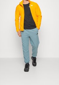 Chaqueta de forro polar naranja, camiseta negra, pantalones de chándal azul claro y zapatillas negras. La chaqueta tiene un cuello alto y cierre de cremallera.