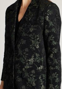 Blazer negro con un sutil patrón floral brillante en verde oscuro, llevado sobre una camiseta negra, mostrando la parte superior del torso y parte del cuello.