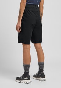 Schwarze Shorts mit glatter Textur, hoher Taille, Seitentaschen und tailliertem Schnitt. Kombiniert mit gemusterten grauen Socken und Schuhen.