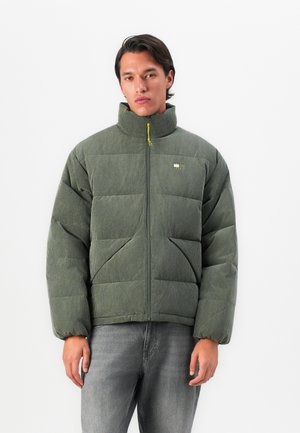 Jeune homme portant une veste matelassée verte à côtes avec un col haut et une fermeture éclair à l'avant, associée à un jean gris, se tenant contre un fond blanc.