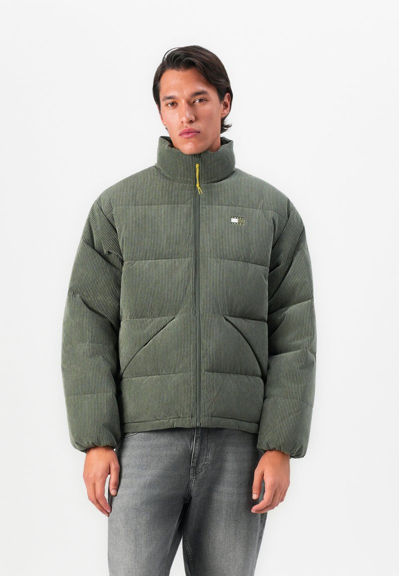 Young man wearing a green ribbed puffer jacket with a high collar and front zipper, paired with gray jeans, standing against a white background.