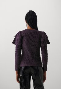 Lila strukturerad stickad topp med volangärmar, med en figurnära silhuett. Kombineras med glänsande svarta byxor, sedd bakifrån.