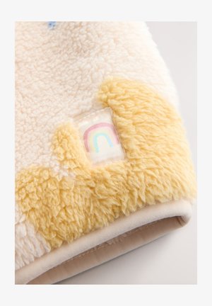 Blød, cremet fleece-tæppe med fluffy gule accenter. Har et lille mærke med et pastelfarvet regnbuedesign. Tekstureret og hyggeligt.