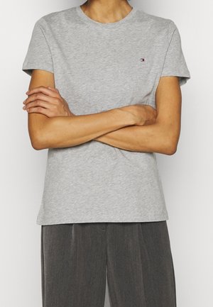 Person iført en lysegrå T-shirt med korte ærmer og et lille logo på brystet, arme krydset, kombineret med mørkegrå bukser.