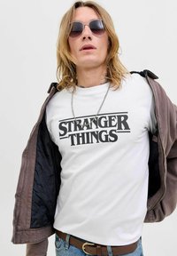 Biała koszulka z napisem "STRANGER THINGS" wydrukowanym dużymi czarnymi literami, noszona pod brązową kurtką, z okrągłym dekoltem i łańcuszkiem na szyi.