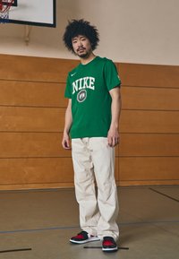 Grøn Nike T-shirt med hvidt logo, beige bukser og sorte/røde sneakers, placeret på en basketballbane med trævæg som baggrund.