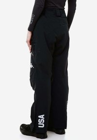 Pantaloni da neve impermeabili neri con ginocchi rinforzati, tasche con zip e un logo sulla coscia. Presentano una vita alta e spalline regolabili.