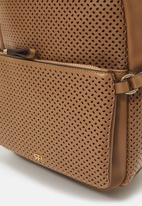 Sac en cuir marron perforé avec fermeture éclair dorée et petit logo doré sur la poche avant, équipé d'une bandoulière latérale avec détail en anneau doré.