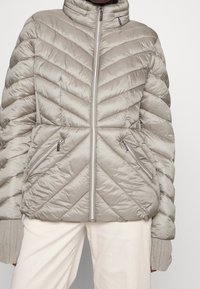 Jasnoszary pikowany puchowy kurtka z wysokim kołnierzem, z charakterystycznym szwem w kształcie zygzaka, z zamykanymi kieszeniami i prążkowanymi mankietami. Gładka, błyszcząca faktura.