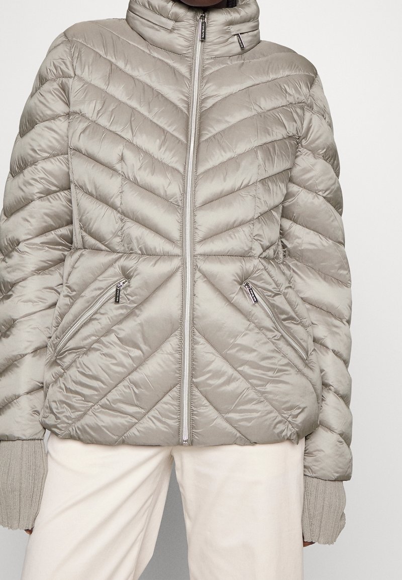 Jasnoszary pikowany puchowy kurtka z wysokim kołnierzem, z charakterystycznym szwem w kształcie zygzaka, z zamykanymi kieszeniami i prążkowanymi mankietami. Gładka, błyszcząca faktura.