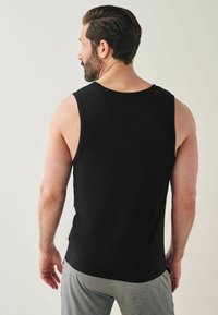 Crna majica na bazi bez rukava od glatkog materijala, s okruglim izrezom i opuštenim krojem. Model nosi svijetlosive hlače.