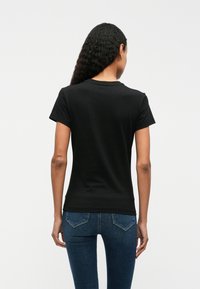 Melna kokvilnas T-kreklā ar īsām piedurknēm un apaļu kakla izgriezumu, redzams no aizmugures. Valkāts ar zilām džinsa biksēm.