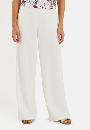 Persona que lleva pantalones blancos de talle alto y pierna ancha con cierre de botón y cremallera, combinados con sandalias doradas de punta abierta y una blusa estampada.