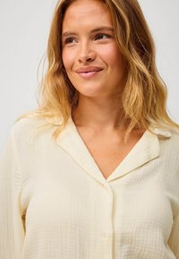 Světle žlutá košile na knoflíky vyrobená z texturované látky, má límec a viditelné záhyby, dlouhé rukávy a volný střih.
