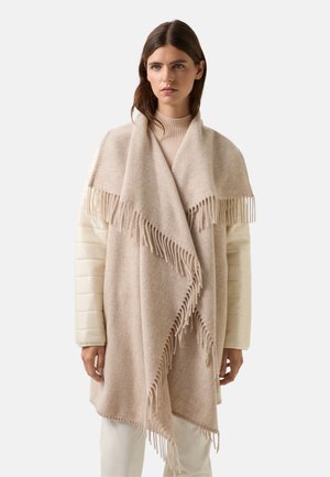 Lys beige poncho lavet af uld, med frynser langs kanterne og en blød tekstur. Bæres over en tætsiddende cremefarvet sweater.