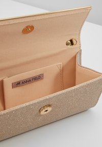 Beige kabelka s texturovaným, třpytivým povrchem. Zlaté kování a západka na zacvaknutí. Interiér podšitý hladkou látkou s kapsou na štítek.