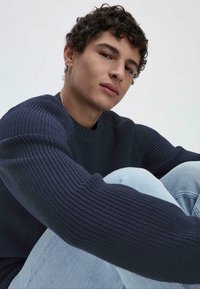 Dunkelblauer Strickpullover mit strukturiertem Rippmuster, rundem Ausschnitt und langen Ärmeln. Hellblaue Jeans mit einem verwehten Farbton.