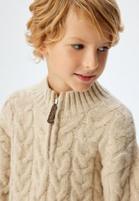 Suéter de punto beige hecho de lana suave, con un alto cuello acanalado y una cremallera corta con un tirador de cuero marrón.