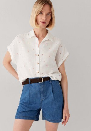 Donna con capelli biondi corti che indossa una camicia bianca a maniche corte con piccoli motivi rossi e pantaloncini di jeans blu con cintura marrone scuro.