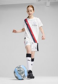 Conjunto de fútbol blanco con una franja diagonal roja y azul marino, con un logo en el pecho. Botas negras y medias blancas hasta la rodilla con acentos.