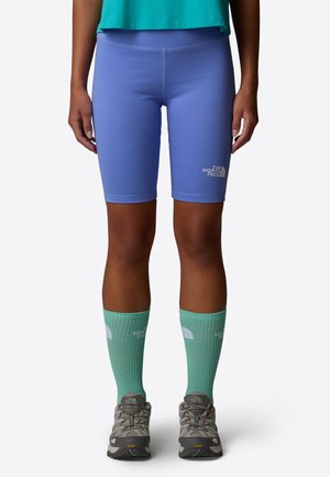 Persoon met blauwgroene korte broek met het logo van The North Face, lichtgroene kniekousen, grijze wandelschoenen en een blauwgroen shirt tegen een witte achtergrond.