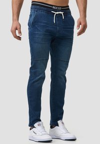 Pantalones jogger de mezclilla azul con cinturilla elástica, cordón y bolsillos laterales. Presentan piernas ajustadas y detalles sutiles en las costuras.