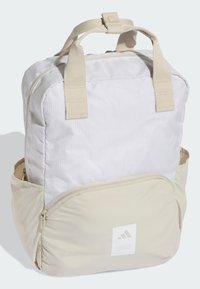Mochila beige y gris claro con textura de cuadrícula, bolsillo frontal con cremallera, asas superiores y correas ajustables. Incluye parche con logo en la parte inferior.
