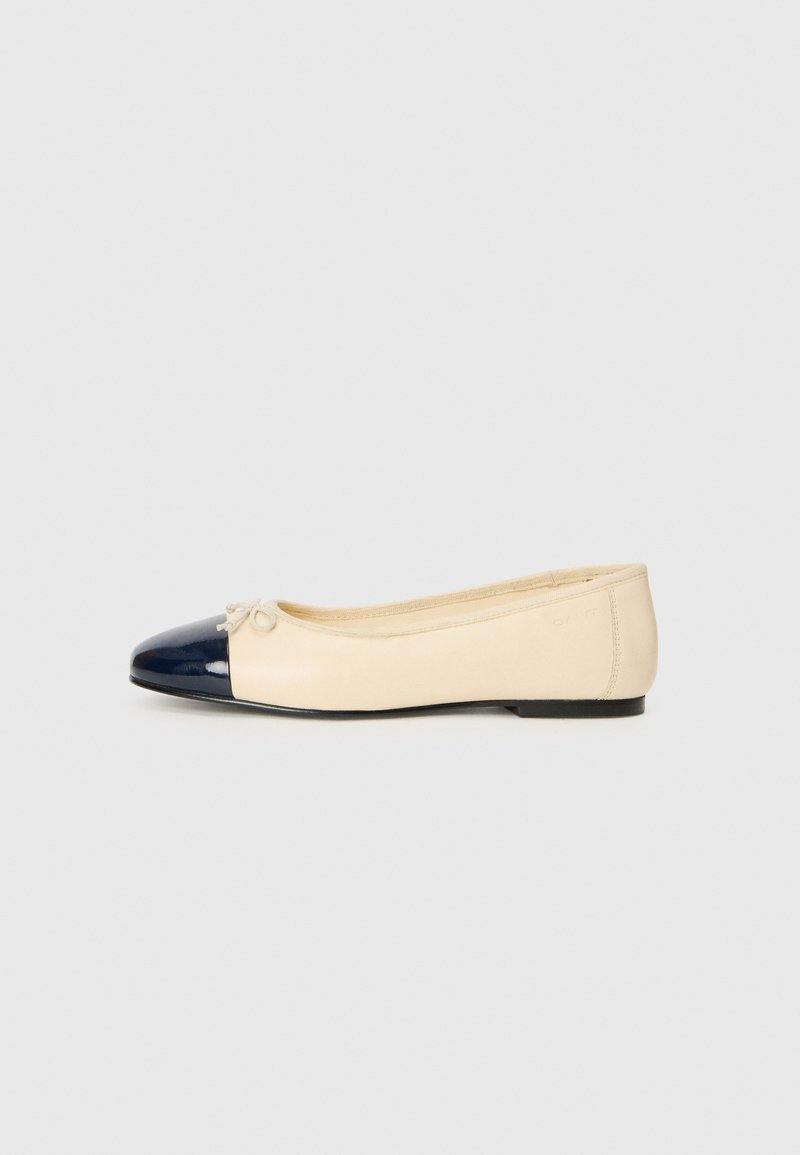 Beige läderballerinor med en glänsande marinblå tåkupp, liten rosettdetalj på framfoten och platt sula, designade för vardagsbruk.