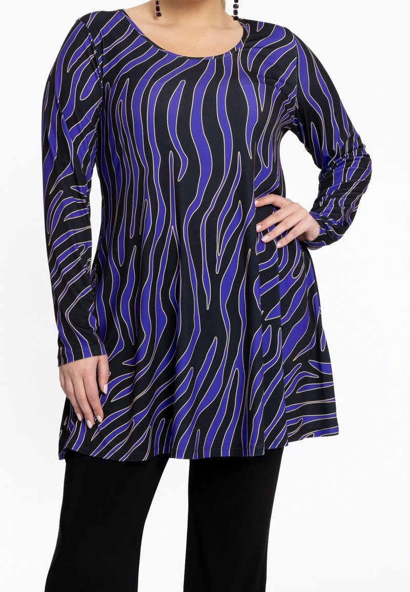 Femme portant une tunique à manches longues rayée zèbre noir et violet avec un pantalon noir, main sur la hanche, sur fond blanc.