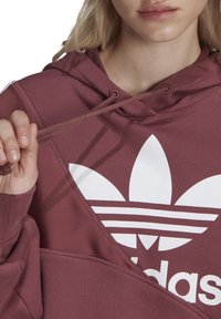 Sudadera con capucha de color burdeos que presenta un gran logotipo blanco de Adidas con tres rayas. Incluye un cordón y una tela texturizada.