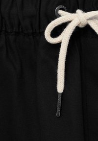 Tissu noir avec une taille élastiquée et un cordon blanc noué en nœud, avec des embouts métalliques et un petit œillet au-dessus.