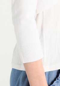 Top de punto blanco con un escote redondo y mangas de tres cuartos con puños acanalados, combinado con un pantalón de mezclilla.