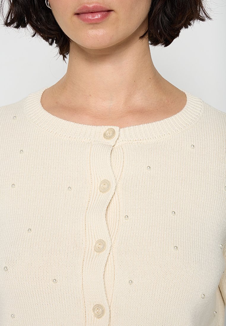 Cremfarbener Cardigan aus strukturiertem Strickstoff, mit kleinen Perlenakzenten und einem runden Ausschnitt mit Knopfverschlüssen.