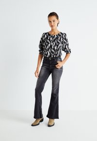 Zwart-witte blouse met een patroon en puffmouwen, gecombineerd met donkere flared jeans en bruine enkellaarsjes, staande tegen een witte achtergrond.
