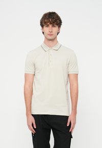 Mladý muž s kučeravými vlasmi, oble�čený v svetlo béžovej polokošeli s krátkym rukávom a čiernych nohaviciach, stojaci na bielom pozadí.