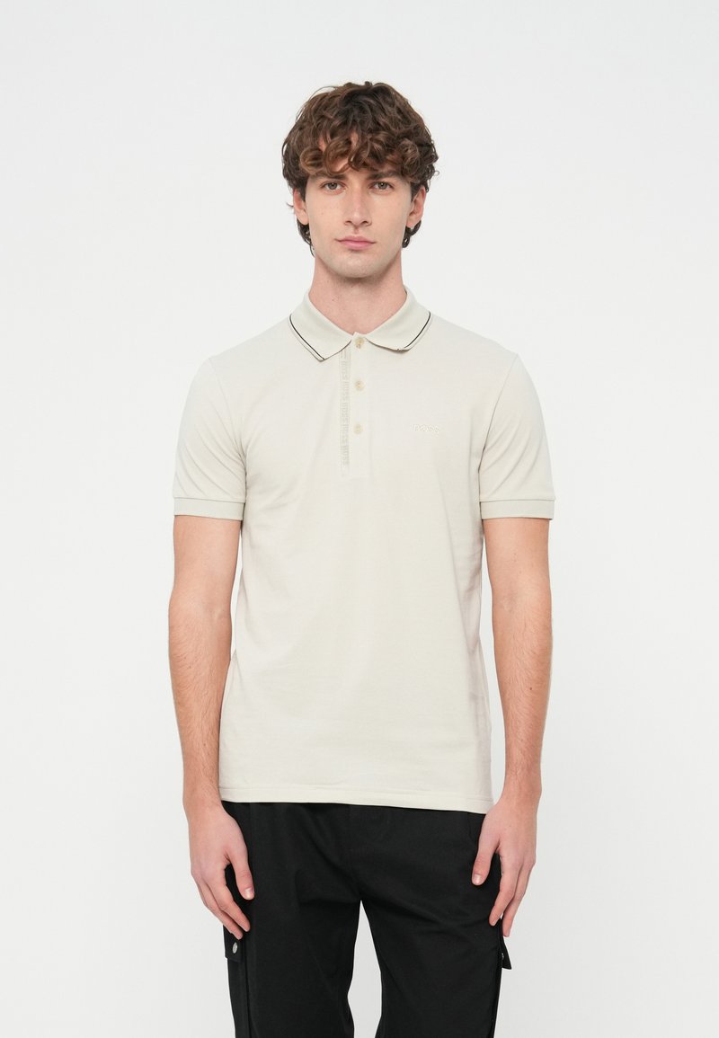 Mladý muž s kučeravými vlasmi, oblečený v svetlo béžovej polokošeli s krátkym rukávom a čiernych nohaviciach, stojaci na bielom pozadí.