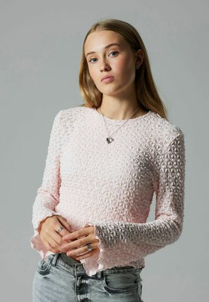 Rosa Langarmoberteil mit strukturiertem Blumenmuster und Rüschen an den Ärmelbündchen, kombiniert mit hellen Denim-Jeans. Silberne Halskette und Ringe sichtbar.