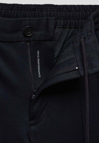 Gros plan sur un pantalon noir avec taille élastique, braguette zippée, fermeture par bouton, cordon de serrage et texte "MNG PERFORMANCE" à l'intérieur de la ceinture.